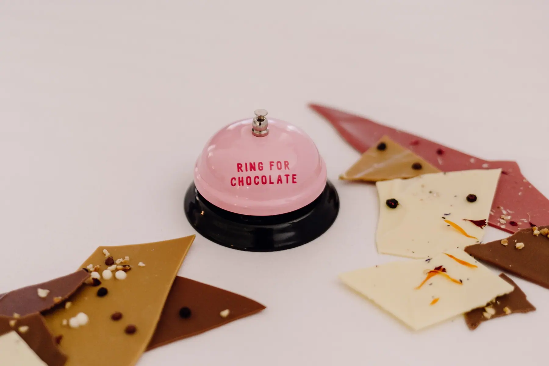 Rosa Tischglocke mit der Aufschrift „Ring for Chocolate“ zwischen Stücken von Bruchschokolade