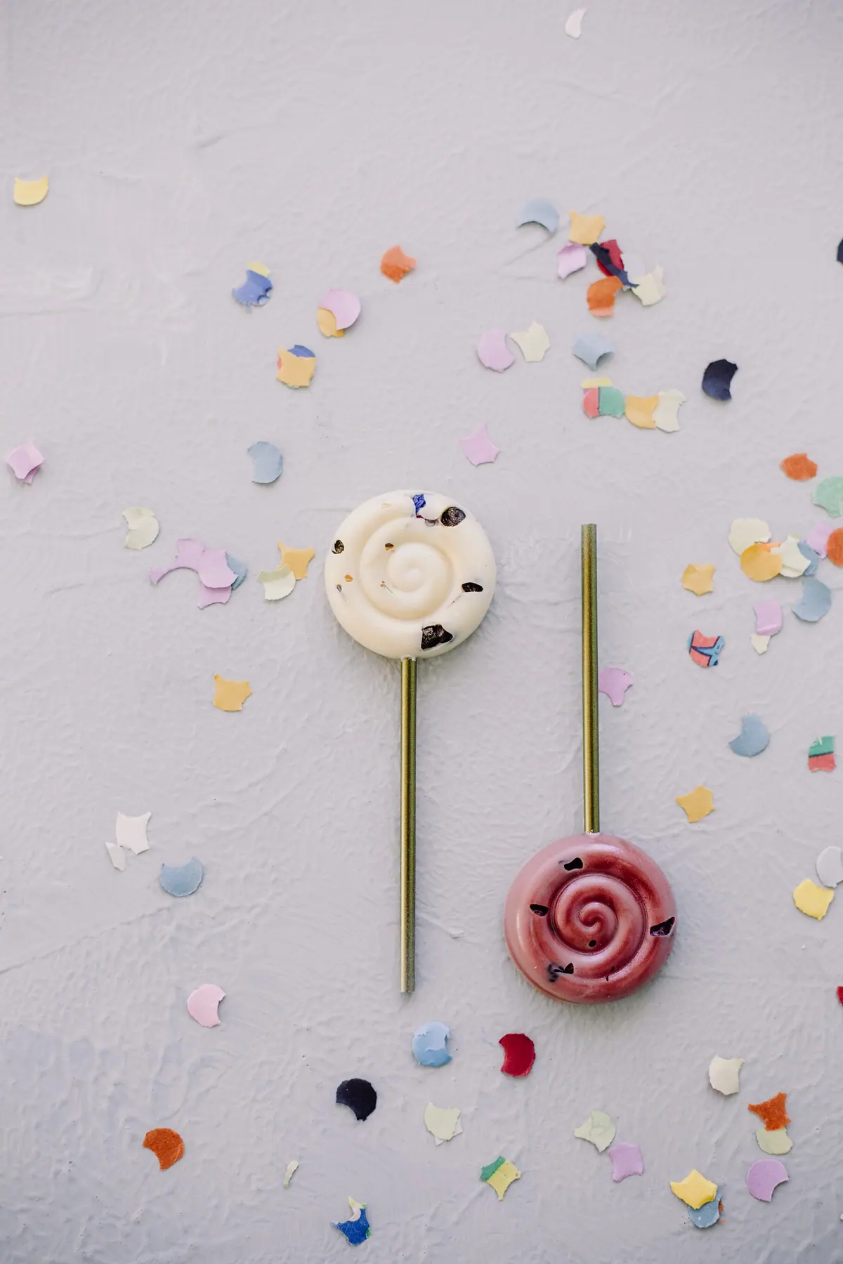 Zwei Schoko-Lollies in Weiß und Rosa an goldenen Stielen auf Konfetti