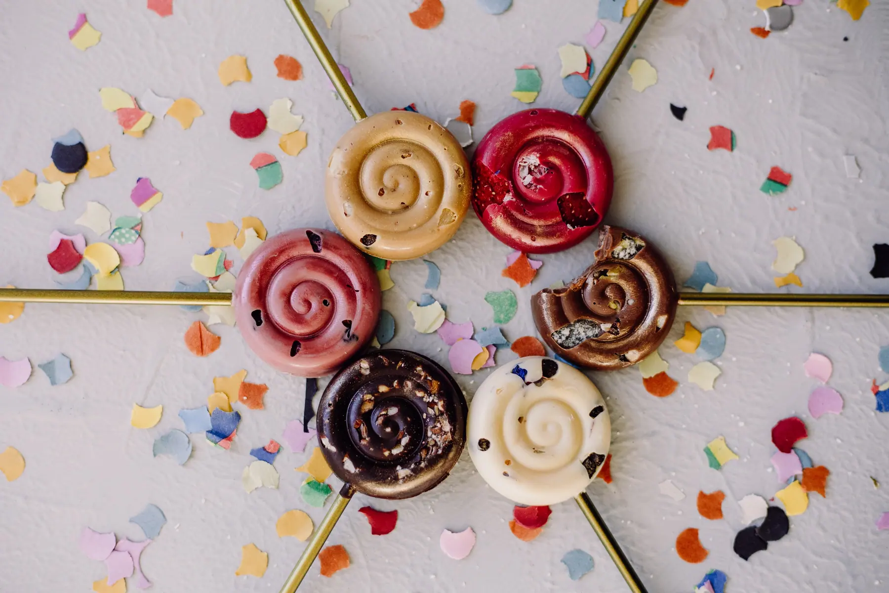 Schoko-Lollies in Schneckenform in Rosa, Weiß und Zartbitter, sternförmig auf Konfetti gelegt