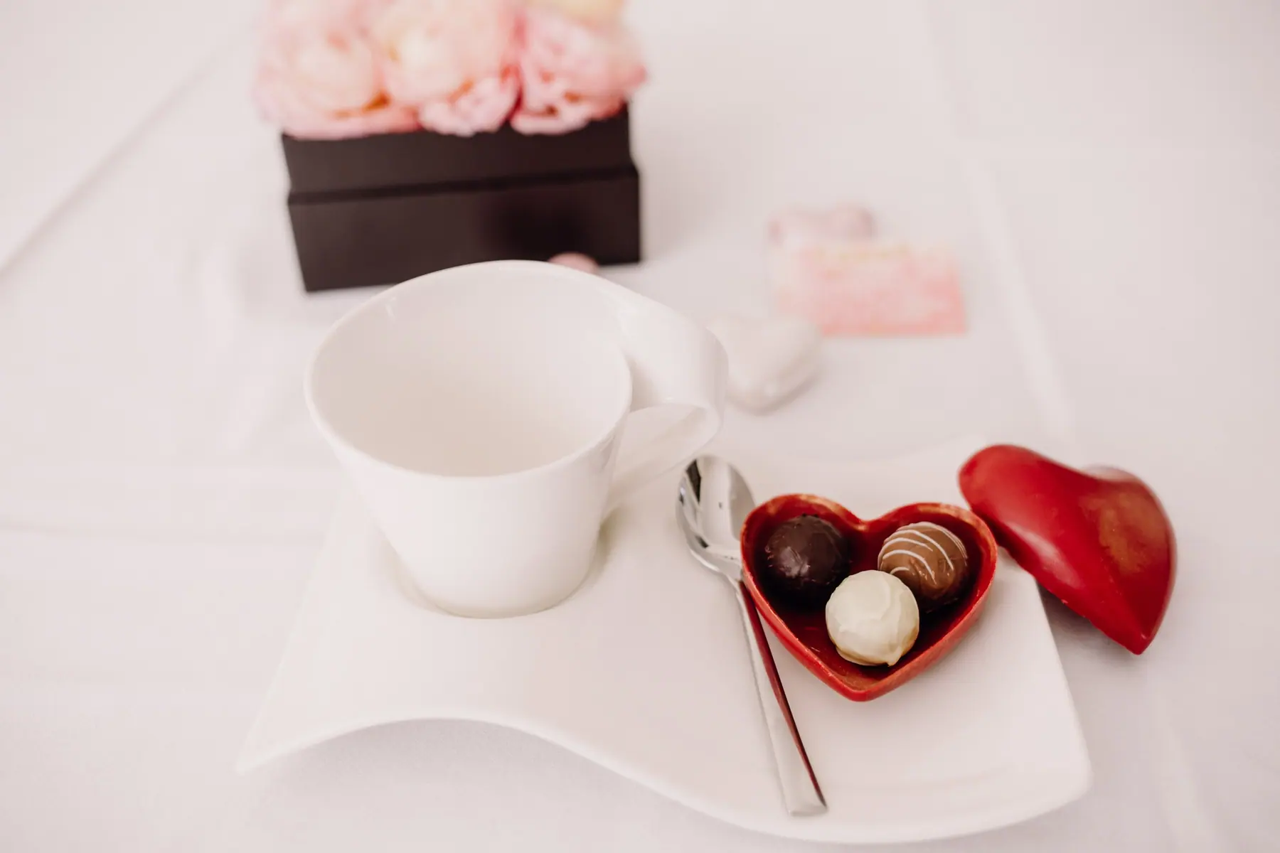 Weiße Tasse auf einer Untertasse mit kleinem Herz voller Pralinen, dahinter eine Rosenschachtel