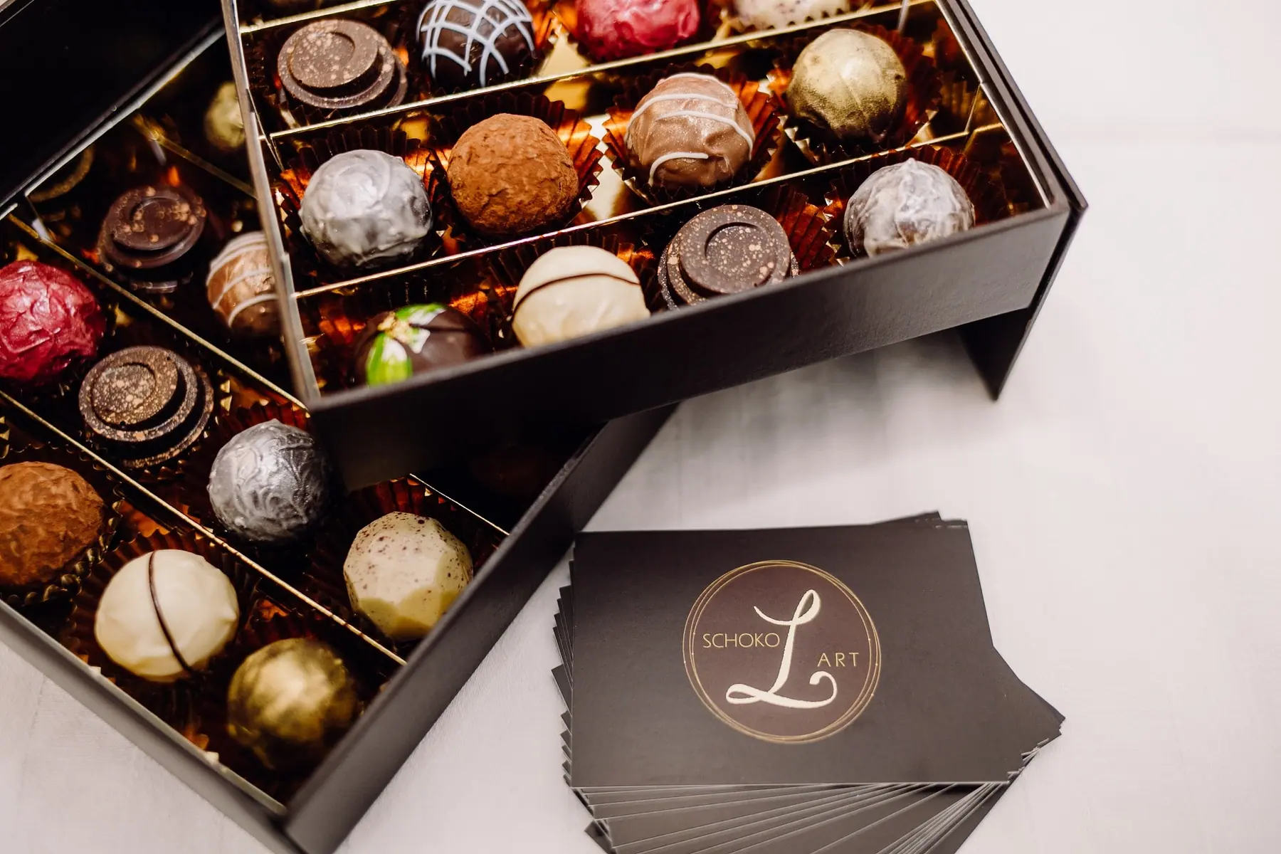 Offene Schachtel mit 32 Pralinen und Trüffeln in Gold, Rot, Weiß und Zartbitter, daneben Visitenkarten mit dem schokoLart-Logo
